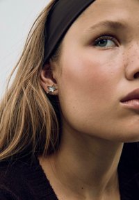 Sølv, stjerneformet ørepynt med en poleret finish, vist på et øre. Brun hårbånd og naturlig hudtekstur i nærbillede.