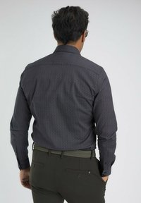 Homme portant une chemise à manches longues à motif sombre, un pantalon vert olive et une ceinture tressée, se tenant avec les mains dans les poches, de dos sur un fond uni.