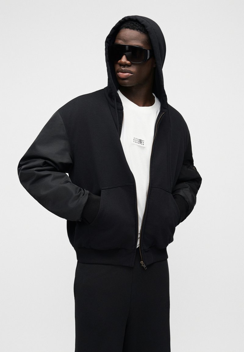 Sort hættetrøje med lynlås, stor hætte, lange ærmer og sidelommer. Båret over en hvid t-shirt med et lille grafikmotiv. Glat tekstur.