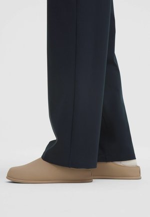 Osoba v béžových slip-on topánkach s bielymi ponožkami a čiernymi širokými nohavicami na jednofarebnom svetlom pozadí.