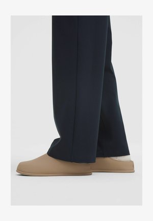 Osoba v béžových slip-on topánkach s bielymi ponožkami a čiernymi širokými nohavicami na jednofarebnom svetlom pozadí.