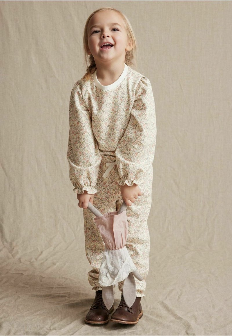 Bloemenprint jumpsuit met lange mouwen en gerimpelde manchetten, met een strik taille en een roze en witte accessoire. Bruine schoenen maken de outfit compleet.