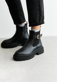 Botas de cuero negras hasta el tobillo con paneles laterales elásticos, suela gruesa y un acento decorativo de hebilla dorada. Llevadas con jeans negros.