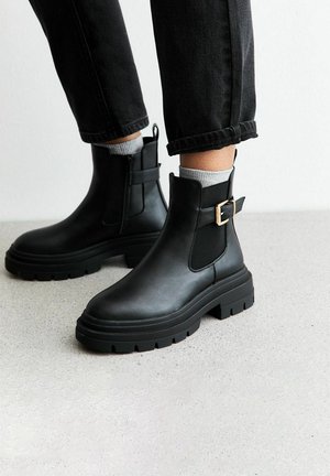 Botas de cuero negras hasta el tobillo con paneles laterales elásticos, suela gruesa y un acento decorativo de hebilla dorada. Llevadas con jeans negros.