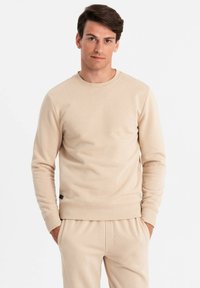 Beige Crewneck Sweatshirt aus weichem Baumwollmix. Mit gerippten Bündchen und Saum, und einem kleinen schwarzen Etikett an der Seite. Sanfte Textur.