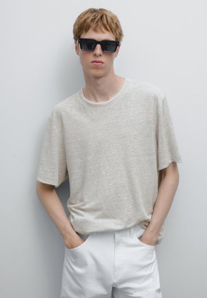 Beige short-sleeve t-shirt with a relaxed fit, paired with white pants. The model wears black rectangular sunglasses, hands in pockets.