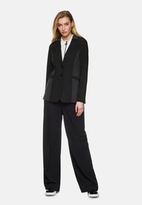 Schwarzer Blazer mit zwei vorderen Knöpfen und kontrastierenden strukturierten Einsätzen. Kombiniert mit lockeren schwarzen Hosen und schwarzen Sneakers.