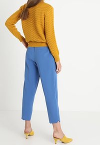 Senfgelber gestrickter Pullover mit einem Zickzackmuster, kombiniert mit blauen, taillierten Knöchellangen Hosen und gelben Absatz-Mules.