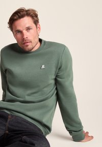 Man with light brown hair wearing a green sweatshirt and dark jeans, sitting with one arm resting behind on a beige background.