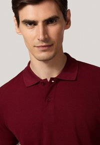 Burgundowa koszulka polo z kołnierzykiem, dwiema guzikami i wyczuwalną tkaniną. Zbliżenie, które podkreśla szczegóły przeszyć.