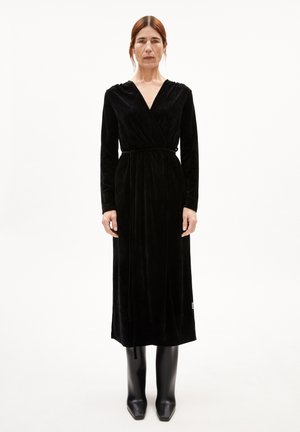 Langes schwarzes Samtkleid mit V-Ausschnitt und tailliertem Bund. Verfügt über lange Ärmel und einen leicht ausgestellten Rock. Getragen mit enganliegenden schwarzen Stiefeln.