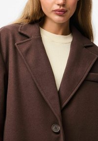 Cappotto in misto lana marrone con colletto a rever, dotato di chiusura con bottone e una tasca sul petto, indossato sopra un dolcevita a coste color crema.
