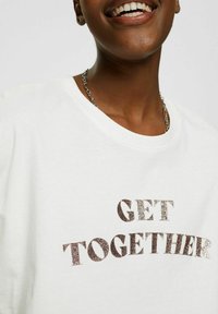 Camiseta de algodón blanca con la frase "UNÁMONOS" en letras metálicas brillantes de color bronce, escote redondo y mangas cortas con un corte relajado.