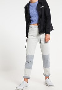 Czarna, lekka kurtka z logo, niebieski, dzianinowy, krótki sweter, szare i białe dżinsy w stylu patchwork oraz białe sneakersy.