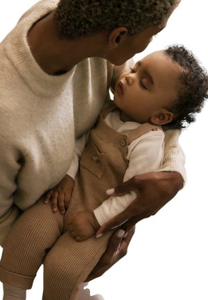 Adulto sosteniendo tiernamente y mirando hacia abajo a un bebé que duerme plácidamente, vestido con un mono de punto beige y una camisa blanca.