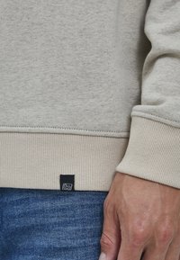 Světle šedá mikina s žebrovanými manžetami a lemem, vyrobená z měkké tkaniny s malou černou cedulkou na spodním lemu.