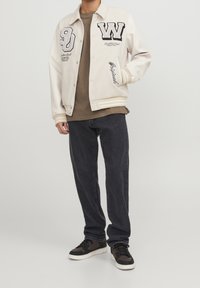 Bomber bunda v krémové barvě s černým nápisem, žebrovanými manžetami a přední kapsou. Nositelná přes hnědou košili a kombinovaná s tmavými džínami a teniskami.