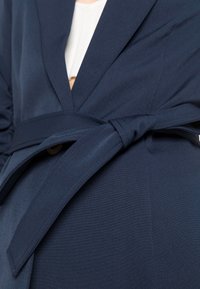 Blazer bleu marine avec taille cintrée par une ceinture, présentant un décolleté en V, un tissu légèrement texturé et une fermeture à un bouton. Design lisse et structuré.