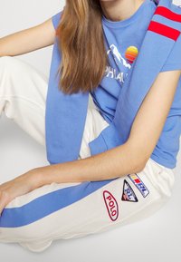 Modrá tišpánka s grafikou hor, bílé tepláky se modrými a červenými pruhy a nášivkami, texturovaná tkanina, volný střih.