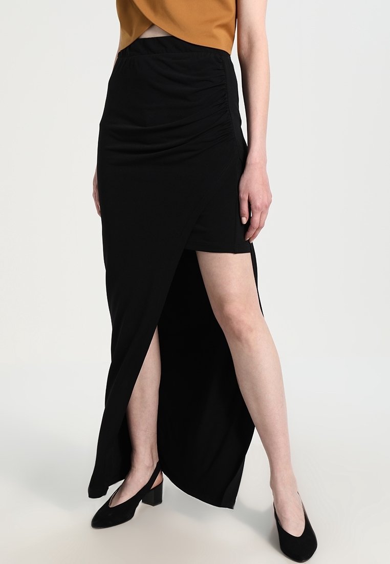 Femme portant une jupe asymétrique noire avec une fente haute sur la jambe gauche, associée à des escarpins slingbacks noirs et un top court marron.