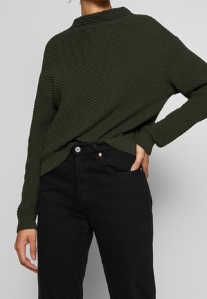 Kvinde iført mørkegrøn tekstureret striktrøje, der er stoppet ind i højtaljede sorte jeans, med den ene hånd i lommen og den anden afslappet.