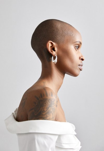 Profilskats uz personu ar cieši noskūtu galvu, tērpušos lielās sudraba auss riņķos un baltā drēbē ar plūstošu audumu.