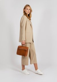 Traje beige a medida con pantalones de pierna ancha, combinado con zapatillas blancas. Sosteniendo un bolso de cuero marrón con un asa superior y detalle de cremallera.