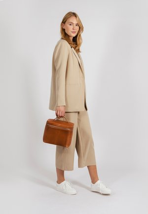 Completo beige su misura con pantaloni a gamba larga, abbinato a sneaker bianche. Tiene in mano una borsa in pelle marrone con manico superiore e zip.