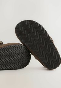 Hnedé semišové topánky s textúrovanými čiernymi gumovými podrážkami s vlnkovým vzorom. Pohľad zozadu, ktorý ukazuje detail podrážky a materiálu.