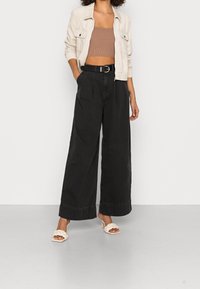 Pantalones anchos negros de cintura alta y con pliegues, combinados con una chaqueta corta beige y sandalias de tacón beige, sobre un fondo neutro.