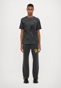 T-shirt noire avec un design graphique saisissant mettant en avant un lion et des motifs floraux. Associé à un pantalon de sweat gris foncé, affichant un logo jaune.