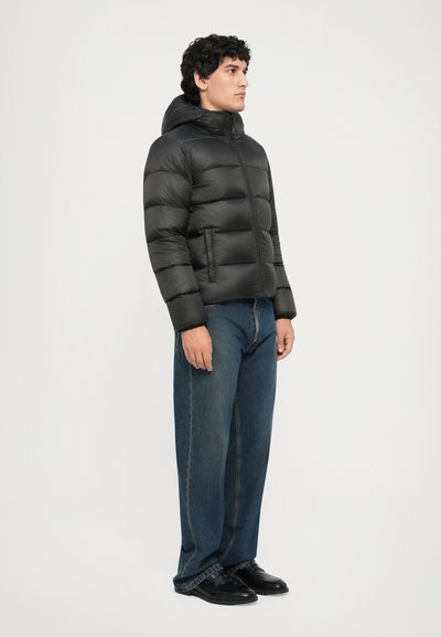 Schwarze Steppjacke mit Reißverschluss, Kapuze und horizontalem Steppmuster; kombiniert mit locker sitzenden blauen Jeans und schwarzen Schuhen.