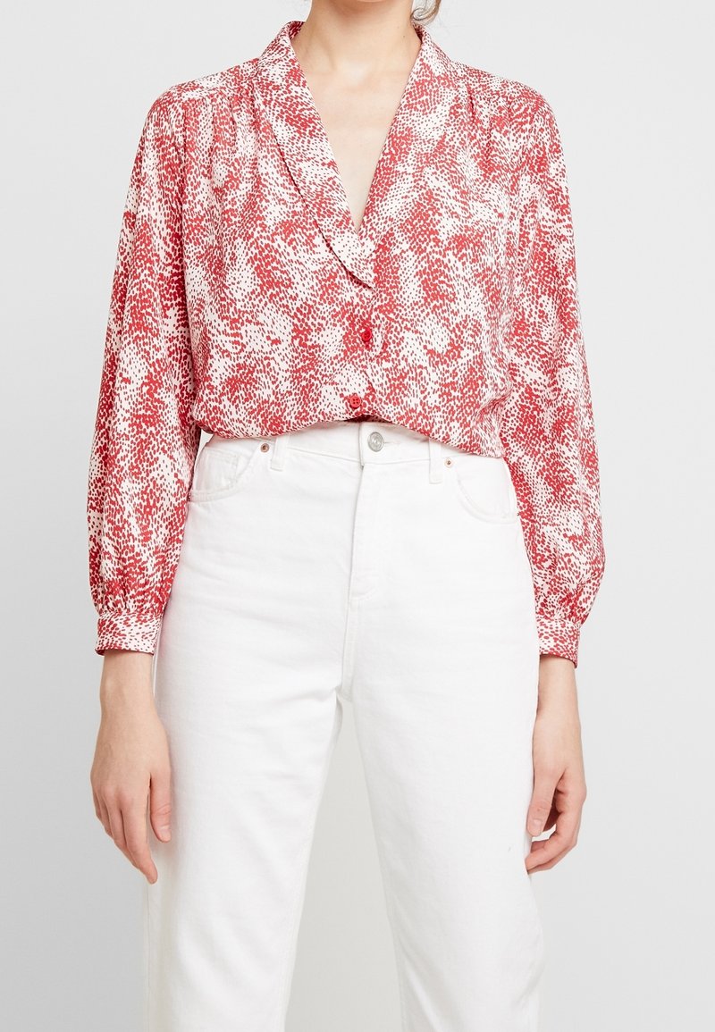 Femme portant un chemisier à manches longues rouge et blanc à motifs, rentré dans un jean blanc taille haute, se tenant devant un fond uni.