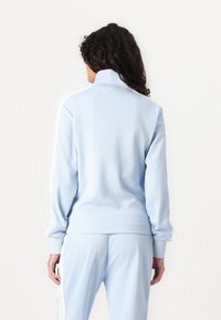 Lys blå zip-jakke med høy krage, ribbede mansjetter og hvite sidestipler på ermene. Myk tekstur, sporty design.