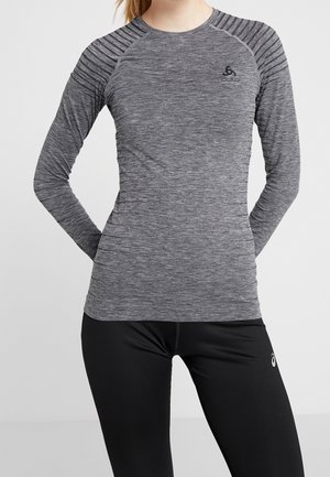 Frau trägt ein eng anliegendes graues meliertes langärmeliges Sportshirt und schwarze Leggings, steht mit den Händen hinter dem Rücken.