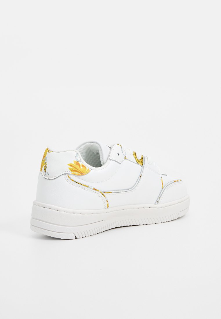 Zapatilla de cuero blanca con un perfil bajo, que presenta acentos en amarillo y plateado metálico, suela de goma texturizada y detalles de diseño minimalista.