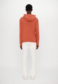 Felpa con cappuccio in arancione bruciato, in misto cotone, con un orlo a costine. Abbinata a pantaloni slim bianchi e sneakers bianche. Vista posteriore.