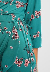 Teal Kleid mit floralen pinken Mustern, Wickeldesign, langen Ärmeln und einem Bindeband in der Taille; aus leichtem Stoff gefertigt.