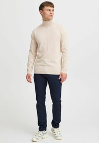 Bež turtleneck pulover z dolgimi rokavi, v kombinaciji z mornarsko modrimi ozkimi hlačami in belimi športnimi copati z modrimi akcenti ter teksturiranim dizajnom.