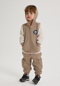 Beige zip hoodie med grå ærmer og et patch. Matchende beige joggerbukser med lommer, båret med sorte sneakers.