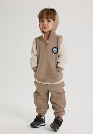 Beige zip hoodie med grå ærmer og et patch. Matchende beige joggerbukser med lommer, båret med sorte sneakers.