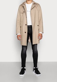 Trench beige con cuello y diseño de doble botonadura, llevado sobre una camisa blanca. Combinado con pantalones vaqueros negros y zapatillas negras.