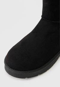 Juodas zomšinis žiemos botas su apvaliu pirštu ir storu guminiu padu šviesiai pilkame fone.