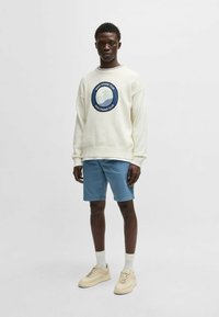 Cream sweater with round blue logo, paired with light blue shorts. Model wears beige sneakers and white crew socks. Neutral background.
