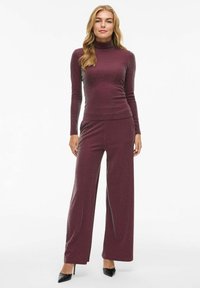 Completo bordeaux con maglione a maniche lunghe e colletto alto abbinato a pantaloni a gamba larga, realizzato in un tessuto testurizzato e luccicante con sottili accenti metallici. Tacchi neri.