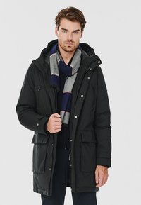 Czarna pikowana parka z kapturem i wieloma przednimi kieszeniami. Noszona na ciemnym swetrze, zestawiona z szaro-granatowym wąskim szalikiem w paski.