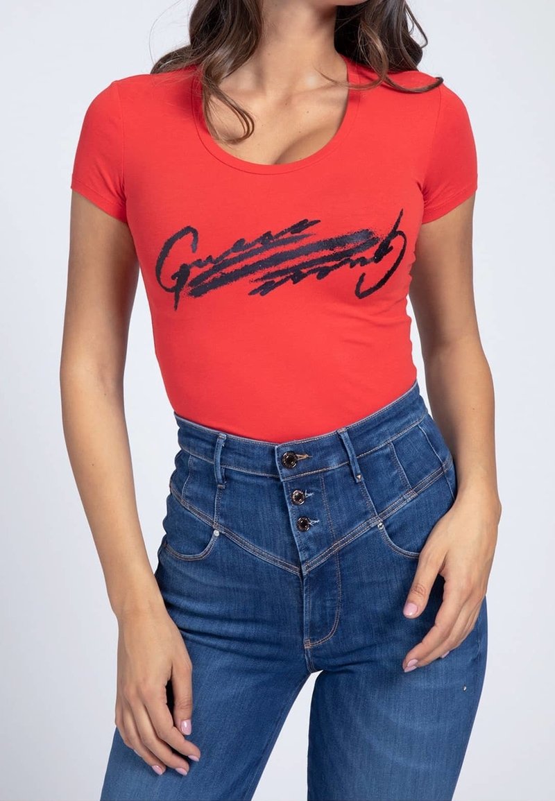 Femme portant un haut rouge ajusté à manches courtes avec un motif en écriture noire, associé à un jean bleu taille haute avec une fermeture à triple bouton.