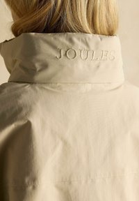 Jachetă ușoară bej cu guler înalt, cu logo-ul brodat "JOULES" pe spate și un material neted, ușor texturat.