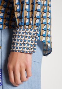 Camicia con motivo geometrico blu, marrone e bianco; presenta polsini con bottoni e dettagli arricciati.