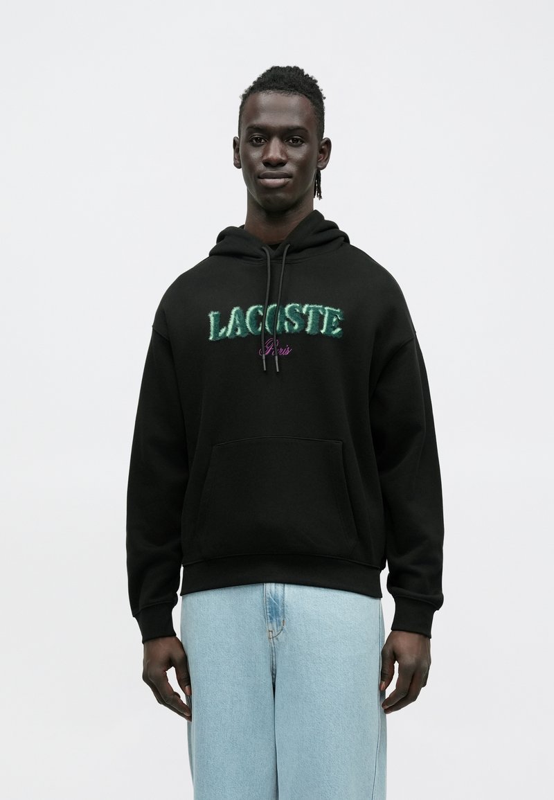 Czarna bluza z kapturem z napisem "LACOSTE" w zielonym kolorze i "Paris" w fioletowym, wyposażona w przednią kieszeń i sznurek do ściągania, noszona z jasnoniebieskimi dżinsami.